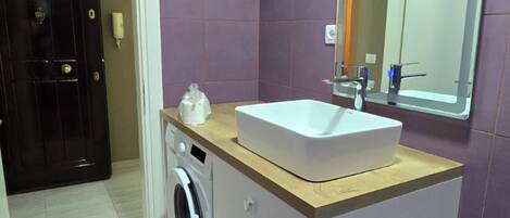 A bathroom with a sink, mirror, washing machine, and a door leading to another room.