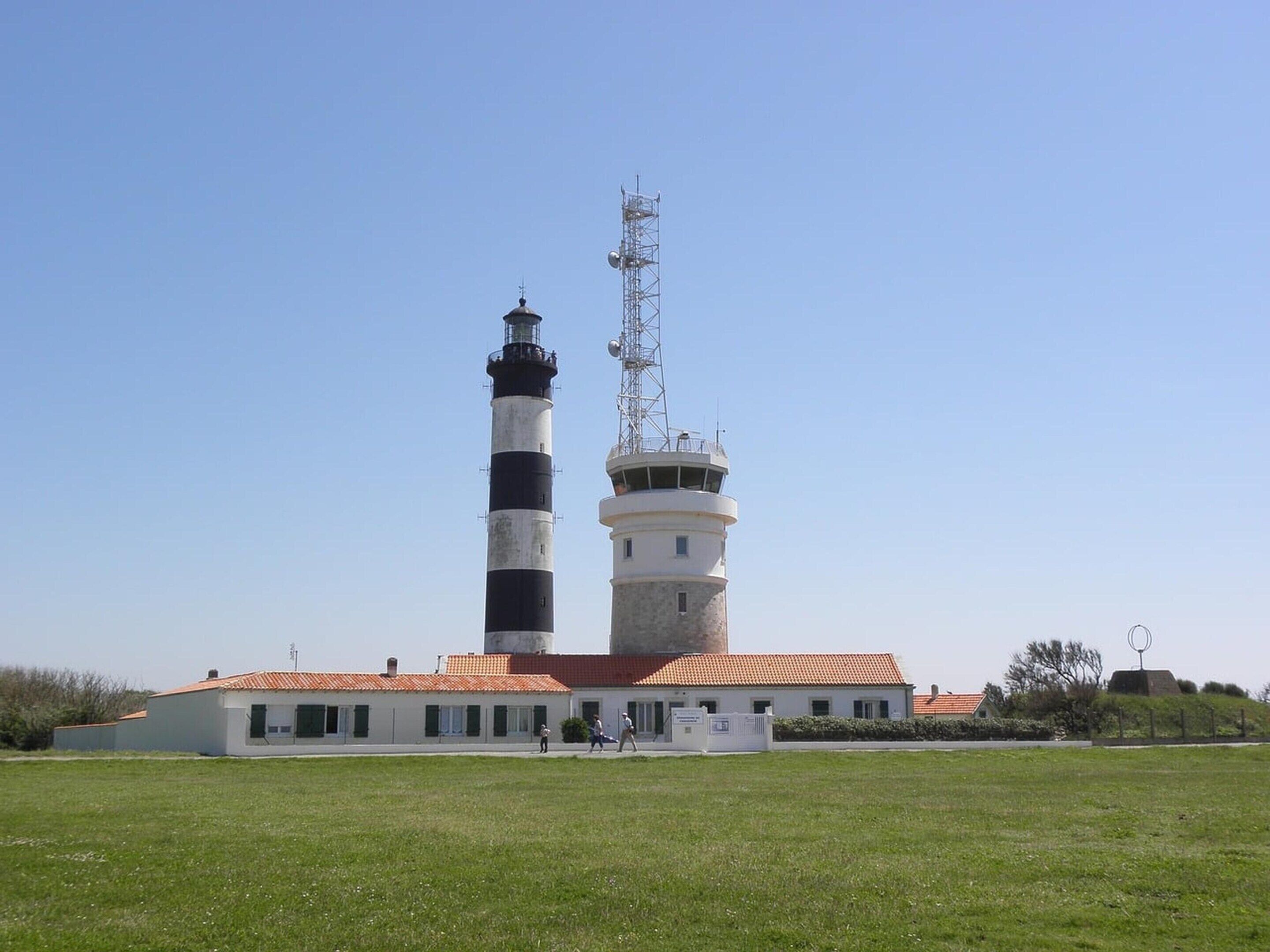 Un faro con una torre a rayas blancas y negras, una torre blanca más pequeña y un edificio con techo de tejas rojas al frente.