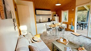 A cozy cabin interior with a sofa, dining table, chairs, a kitchen area, and a sliding glass door leading to an outdoor seating area. - Coastal Family Cottage for Six (Saint-Denis-d'Oleron)