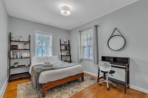 Un dormitorio con una cama, dos mesitas de noche, un escritorio con una silla y dos ventanas con cortinas.