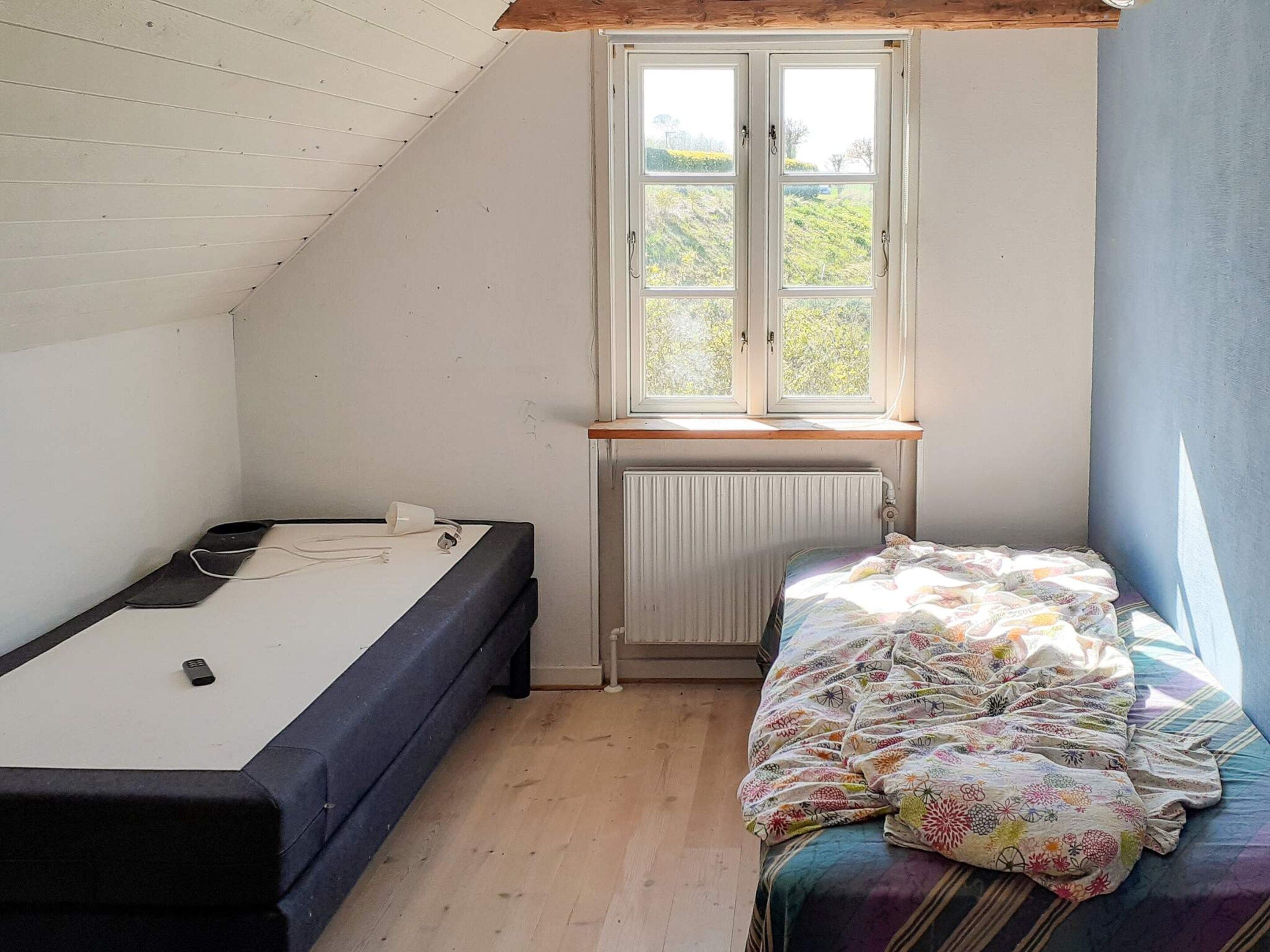 Un alojamiento pequeño y de una sola habitación, con techo inclinado, ventana con vista a follaje, cama con colcha colorida y escritorio con silla.