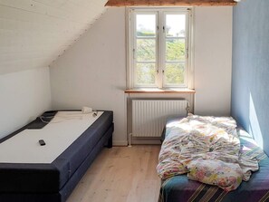 Un alojamiento pequeño y de una sola habitación, con techo inclinado, ventana con vista a follaje, cama con colcha colorida y escritorio con silla.