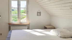 Un dormitorio con una cama grande, una ventana con vista a follaje, una mesita de noche pequeña y un techo inclinado.