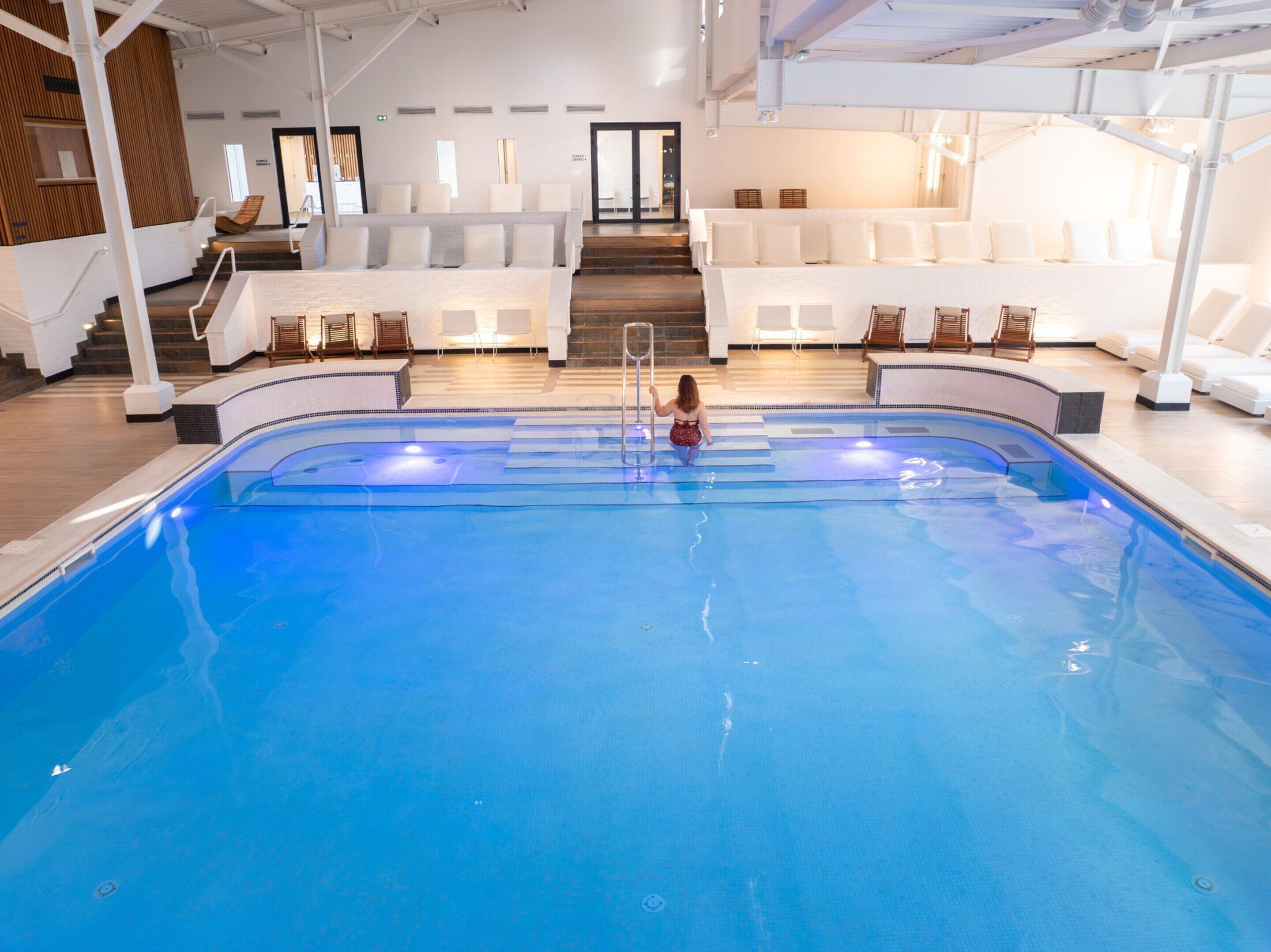 Une piscine intérieure où un enfant nage, entouré de chaises longues blanches et d’un escalier menant à un niveau supérieur.