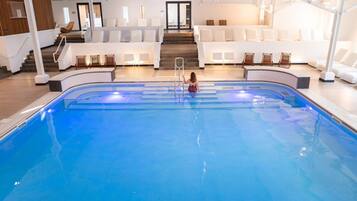Une piscine intérieure où un enfant nage, entouré de chaises longues blanches et d’un escalier menant à un niveau supérieur.