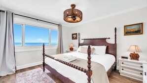 Ein Bett mit Baldachin, ein Holzkopfteil, ein Nachttisch mit Lampe, Blick auf das Meer durch ein Fenster und ein Bild an der Wand.