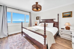 A four-poster bed with a wooden headboard, a bedside table with a lamp, a view of the sea through a window, and a painting on the wall.