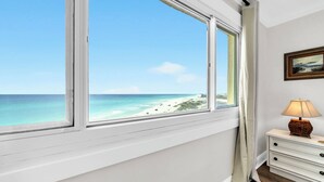 Ein Zimmer mit großem Fenster mit Blick auf einen Strand, einem Nachttisch mit Lampe und einem gerahmten Bild an der Wand.
