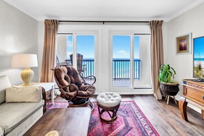 A living room with a sofa, armchair, ottoman, lamp, and a view of the ocean through large windows.