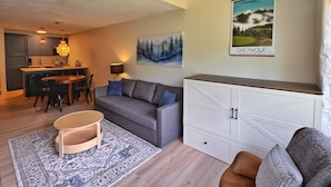 A modern living room with a grey sofa, a small wooden table, a patterned rug, a white cabinet, and a painting of a forest.