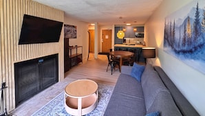 A modern living room with a flat-screen TV, a fireplace, a round coffee table, a sofa, and a dining area with chairs.