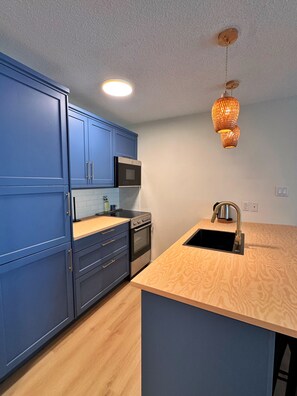 Une cuisine moderne avec des armoires bleues, un plan de travail en bois, un four encastré, un four à micro-ondes et un luminaire suspendu.