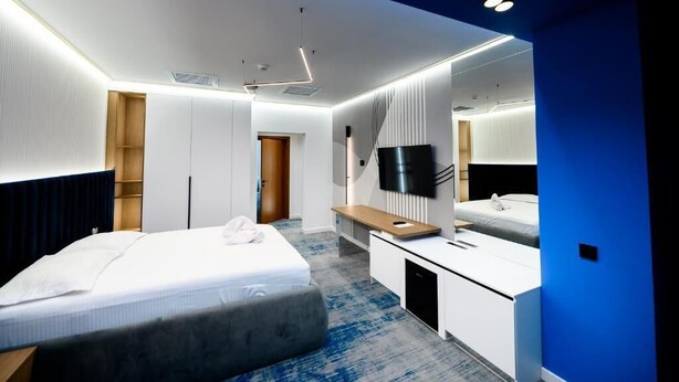 A modern hotel room with a bed, bedside table, TV, and a large mirror. - Hotel Bavaria (Craiova)