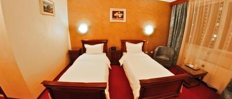 Habitación de hotel con dos camas de madera, alfombra roja, cabeceros de madera, una mesita, una silla y una ventana con cortinas.