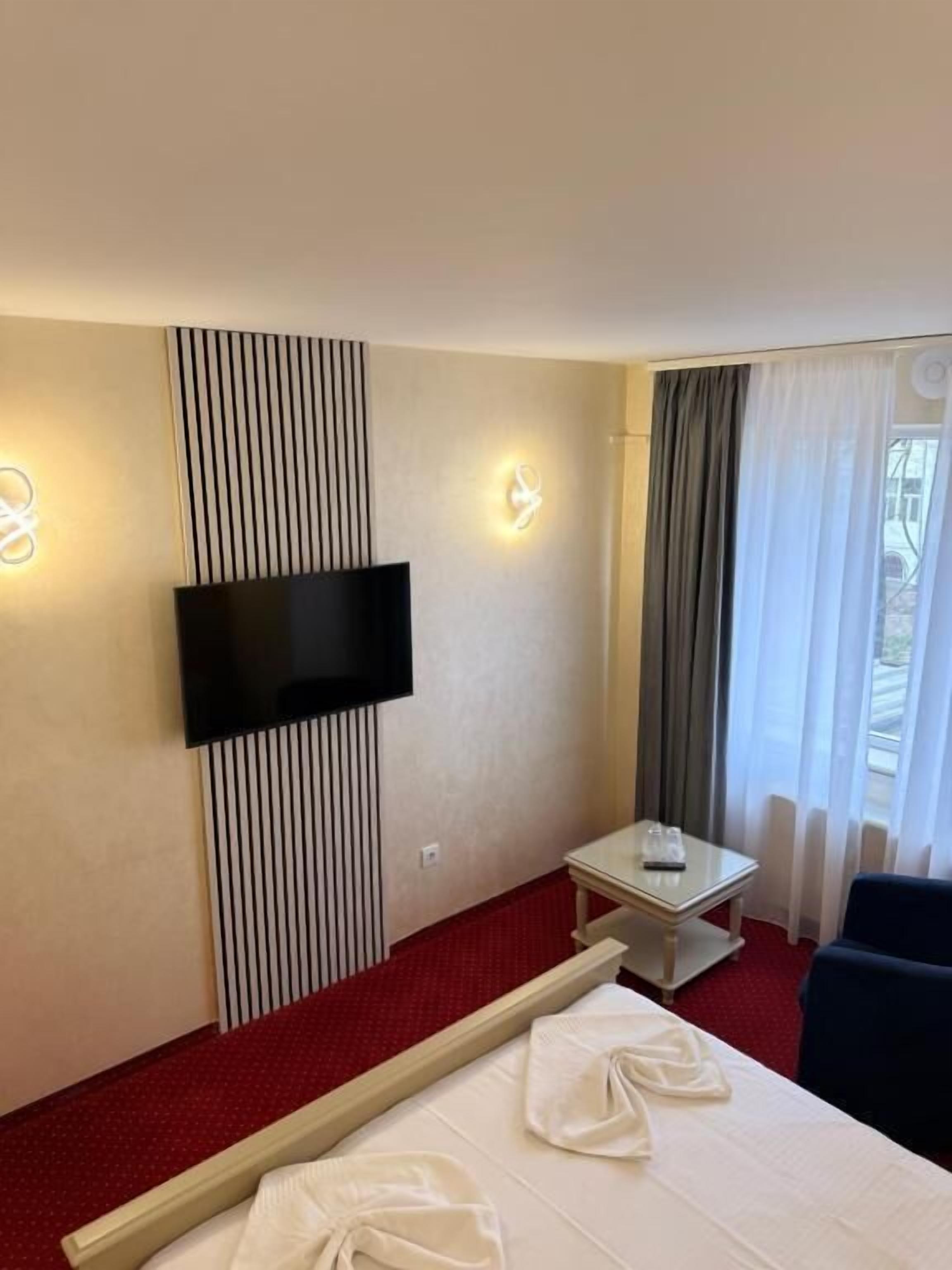 Una habitación de hotel con una cama, televisión instalada en la pared, una mesita y una ventana con cortinas.