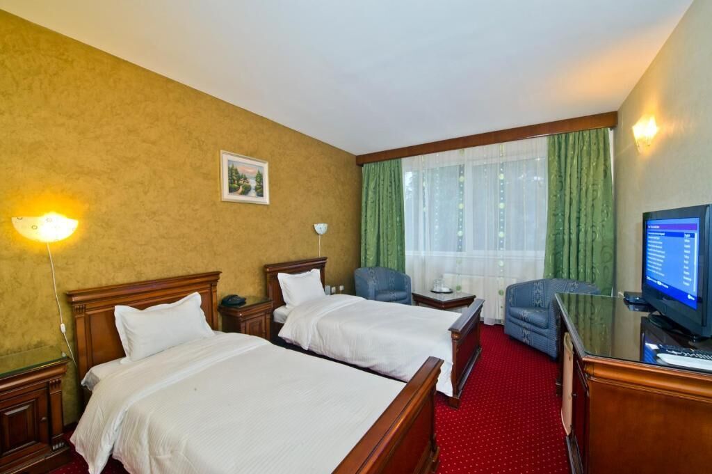 Quarto de hotel com duas camas, uma escrivaninha com computador, uma cadeira, televisão e janela com cortinas.