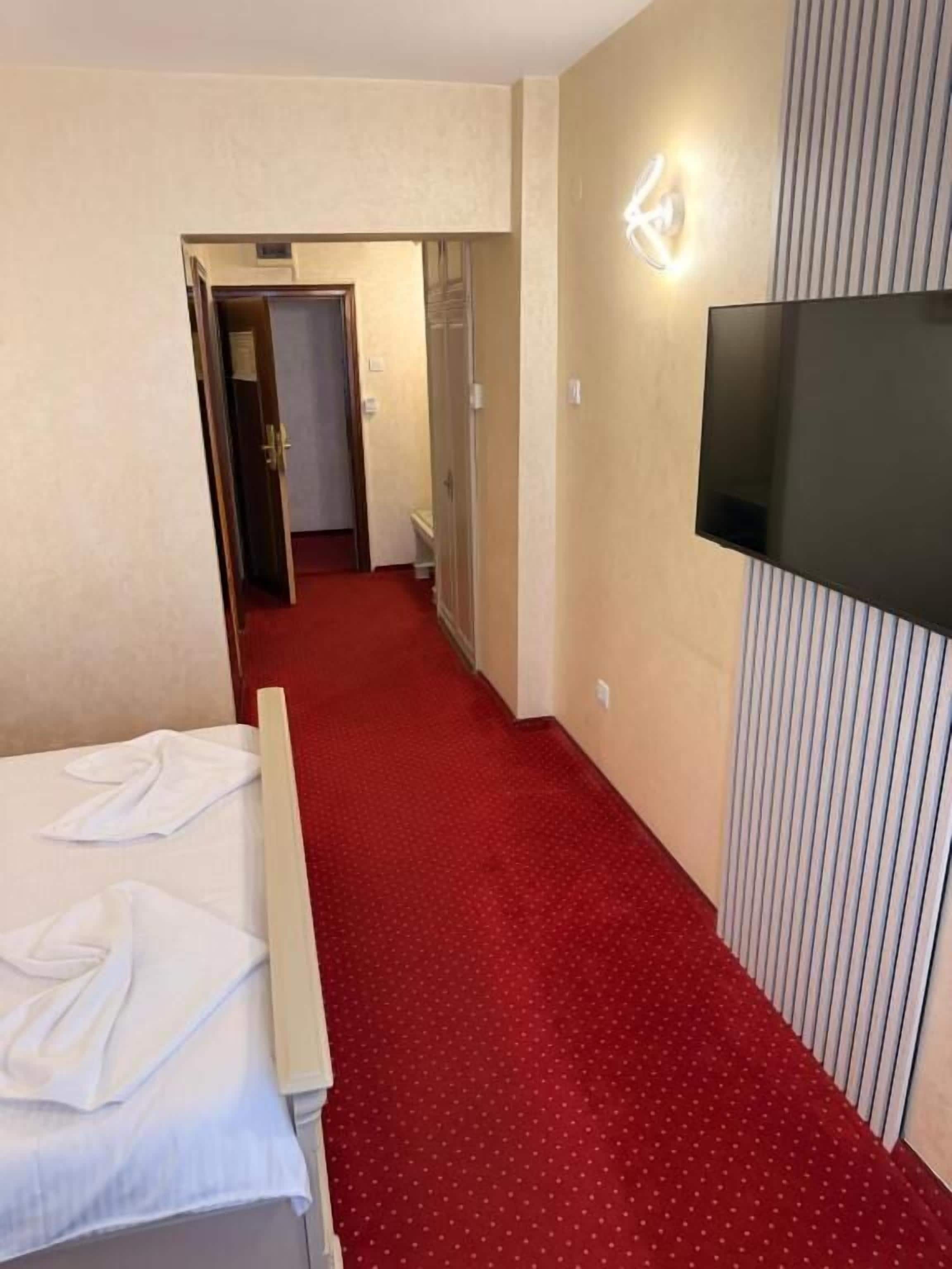 Habitación de hotel con cama, televisor montado en la pared, pasillo con alfombra roja y puerta de acceso al baño.
