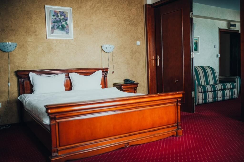 Een hotelkamer met een houten bed, een rood tapijt, een groen gestreepte bank en een deur die naar een andere kamer leidt.