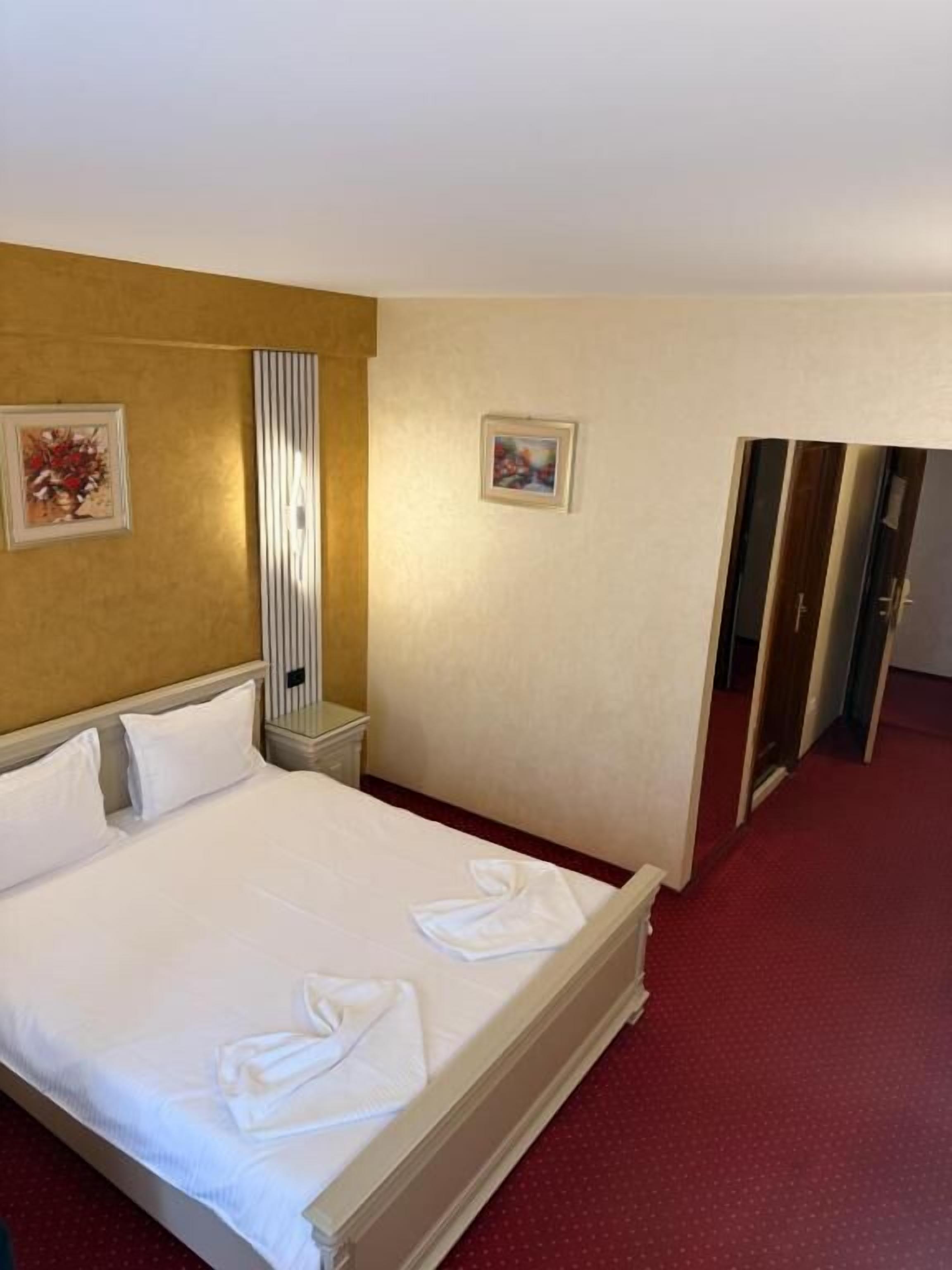 A hotel room with a bed, a nightstand, a wall-mounted lamp, and two framed pictures.