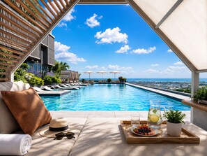 Zona junto a la piscina con sillones de descanso, una tumbona, una bandeja con frutas y una bebida, y vistas al océano y al paisaje urbano.