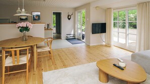 A spacious living area with a wooden dining table, a sofa, a TV mounted on the wall, and large windows offering a view of greenery outside. - 4 Star Holiday Home in Gilleleje (Gilleleje)
