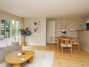 A living room with a sofa, coffee table, dining table, chairs, a potted plant, and a white door leading to another room. - 4 Star Holiday Home in Gilleleje (Gilleleje)