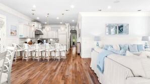 Una cocina amplia con armarios blancos, un comedor con sillas de mimbre y una sala de estar con sofá blanco y cojines azules.