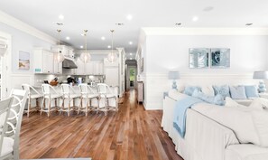 Eine geräumige Küche mit weißen Schränken, ein Essbereich mit Korbstühlen und ein Wohnzimmer mit weißem Sofa und blauen Kissen.