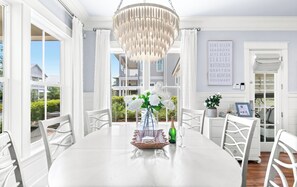 Un comedor con una mesa blanca, sillas, una lámpara de araña y vistas a una casa a través de amplios ventanales.
