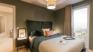 Ein Schlafzimmer mit einem großen Bett, einem Nachttisch mit Lampe, Blick auf die Stadt durch ein Fenster und einer außerhalb liegenden Stadtansicht.