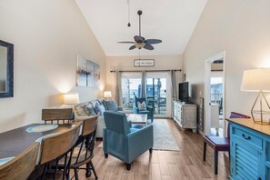 A modern living room with a dining area, a ceiling fan, a TV, and a view of the city. - Little Lagoon + Beach Access | Pool + Fishing Dock | The Landing 102 (Gulf Shores)