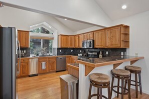 Una cocina moderna con armarios de madera, un refrigerador de acero inoxidable, un microondas y una estufa integrada. Hay taburetes de bar y una ventana que proporciona luz natural.