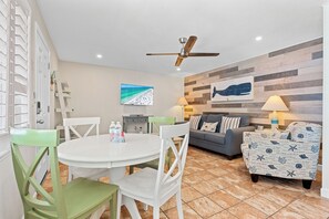Une pièce comprenant une table de salle à manger ronde, des chaises, un canapé, un fauteuil à motifs, un ventilateur de plafond, un téléviseur fixé au mur et une œuvre d’art sur le thème de la plage.