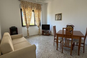 A living room with a sofa, a TV, a wooden dining table with chairs, a window with curtains, and a framed picture on the wall.