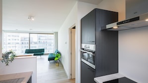A modern kitchen with a built-in oven and a stove, a dining area with a table and chairs, and a view of a cityscape through large windows. - Stunning Central Antwerp Stay for 12 (Antwerp)