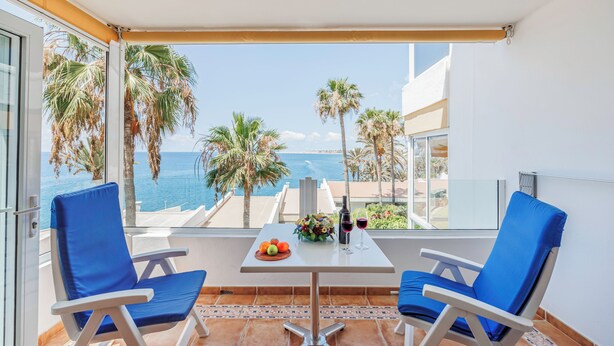 A balcony with two blue chairs, a table with fruit and wine, and a view of the sea and palm trees. - Sunsuites Collection - Las Flores (San Bartolome de Tirajana)