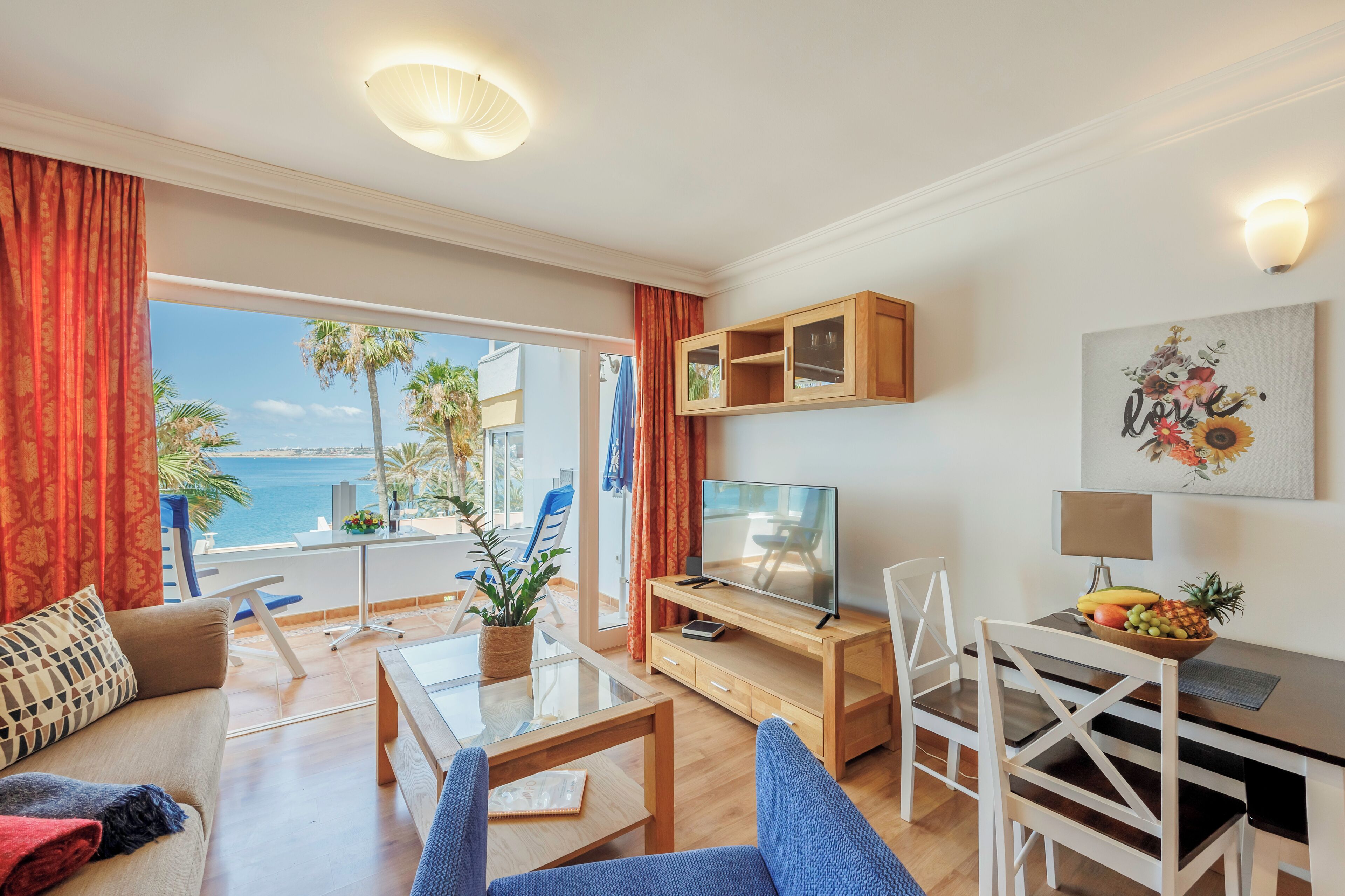 Un salon moderne avec un téléviseur, une table à manger et des chaises. Il y a un balcon avec vue sur la mer.