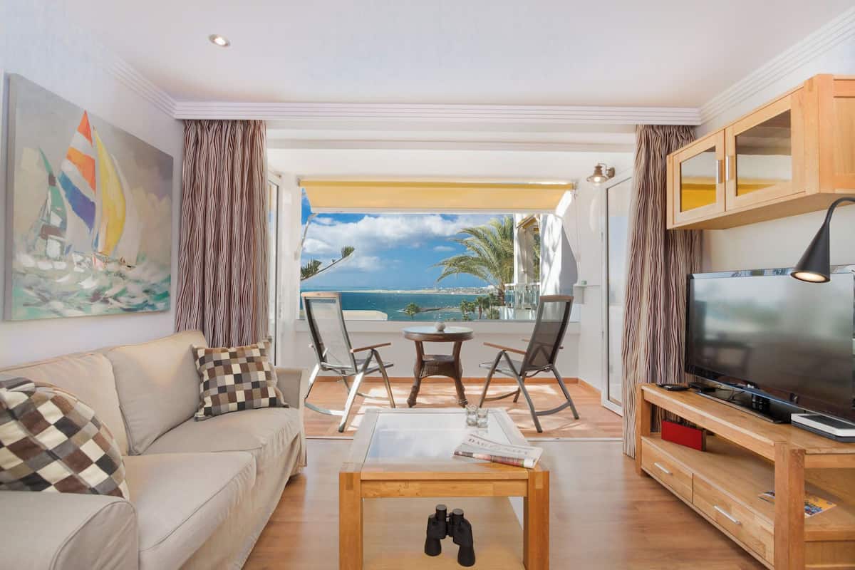 A living room with a sofa, a coffee table, a TV, and a balcony with a view of the sea.