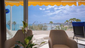 A balcony with two lounge chairs, a table, and a view of the sea and palm trees. - Sunsuites Collection - Las Flores (San Bartolome de Tirajana)