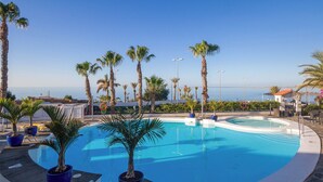 A swimming pool with palm trees, a view of the sea, and a small building in the background. - Sunsuites Collection - Las Flores (San Bartolome de Tirajana)