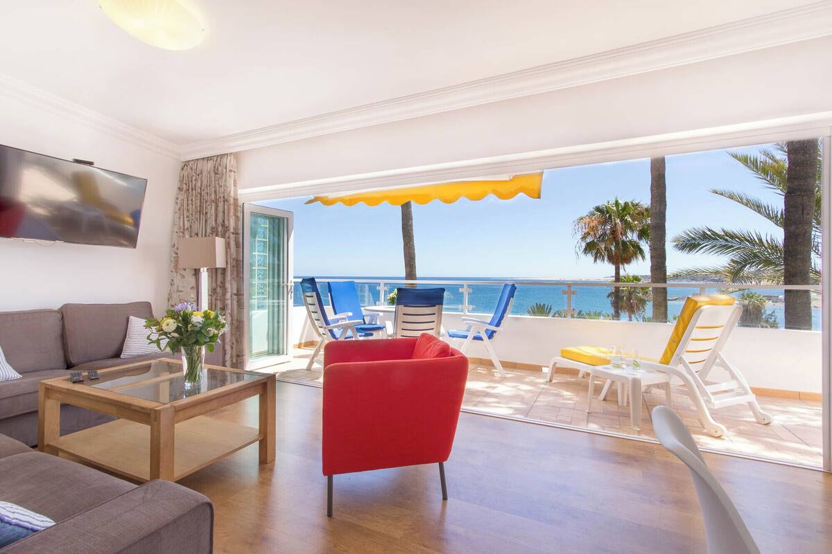 A living room with a sofa, coffee table, and a red chair. There is a balcony with a yellow umbrella, blue and white lounge chairs, and a view of the sea.