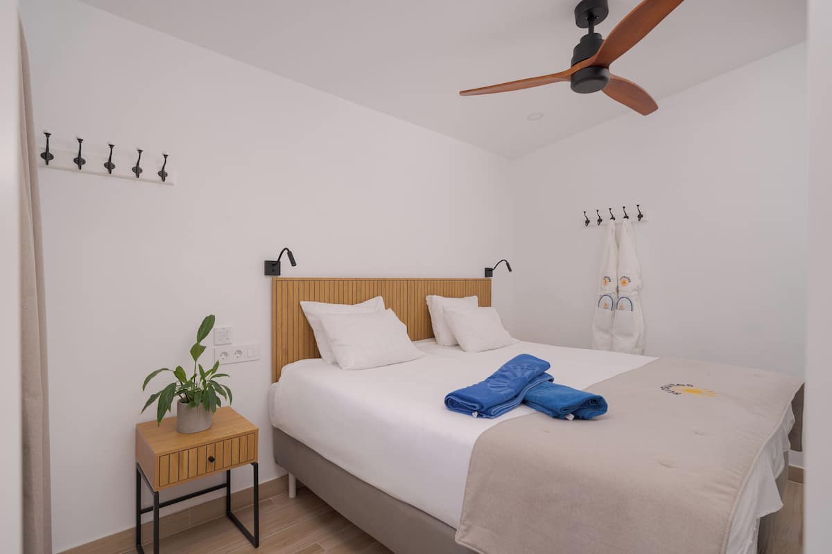 A bedroom with a bed, a ceiling fan, a bedside table with a plant, and a wall-mounted light fixture.