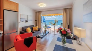 A modern living room with a kitchen, a red chair, a sofa, a coffee table, a lamp, and a view of the ocean. - Sunsuites Collection - Las Flores (San Bartolome de Tirajana)