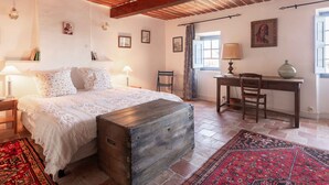 Quarto com cama de madeira, uma escrivaninha, uma cadeira, janela com cortinas e um tapete.