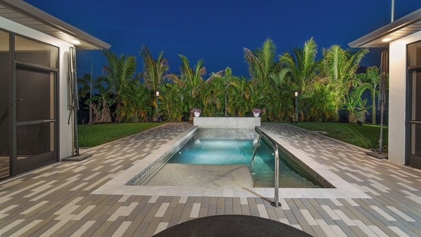 Una piscina con solárium empedrado, rodeada de palmeras y un edificio con puertas de cristal.