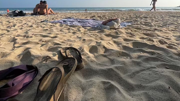 A sandy beach with footprints, a pair of sandals, and a blanket.