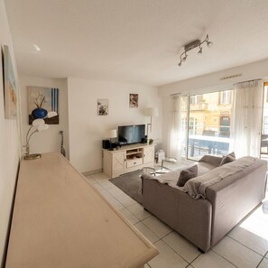 A living room with a sofa, a TV, a lamp, and a window with a view. - Apartment with balcony in the heart of Le Touquet (Le Touquet-Paris-Plage)