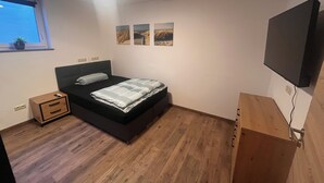Quarto com cama, mesa de cabeceira, televisão na parede e janela com planta.