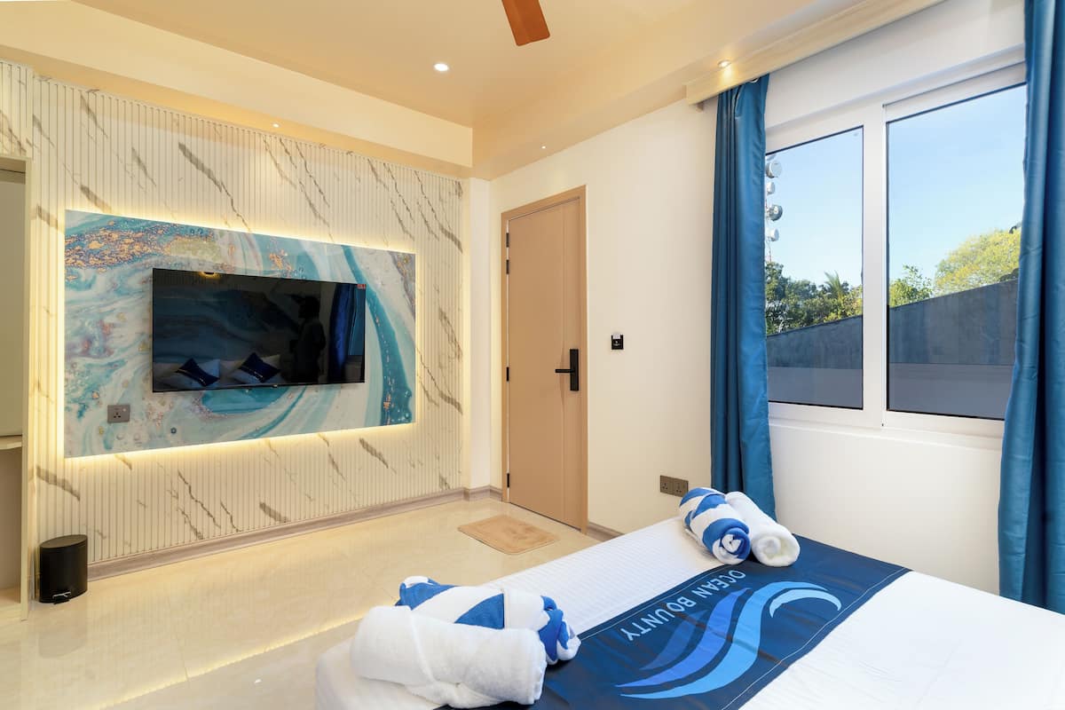 Une chambre d’hôtel moderne avec un grand téléviseur fixé sur un mur en marbre, un lit recouvert d’une courtepointe bleue et blanche, et une vue sur l’extérieur par une fenêtre.