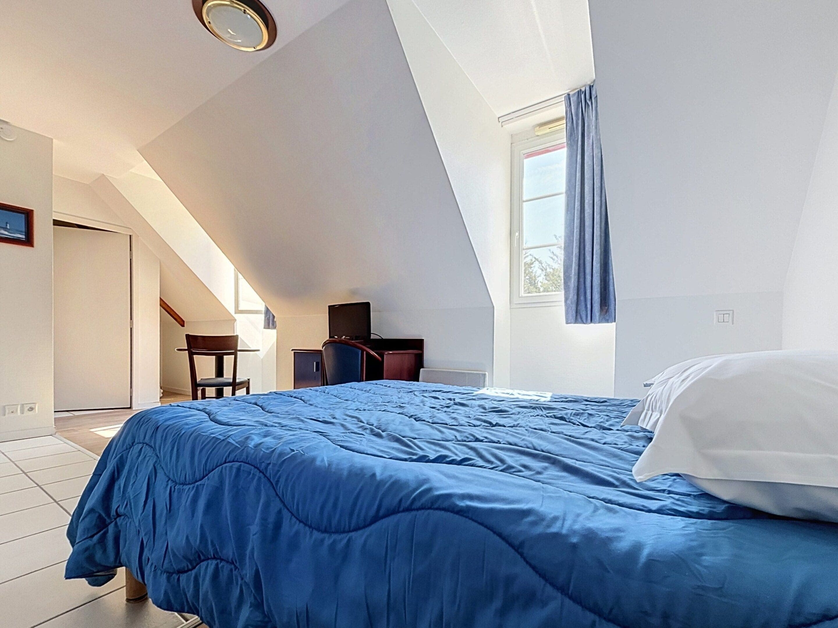 A bedroom with a bed, a desk, a chair, and a window with curtains.